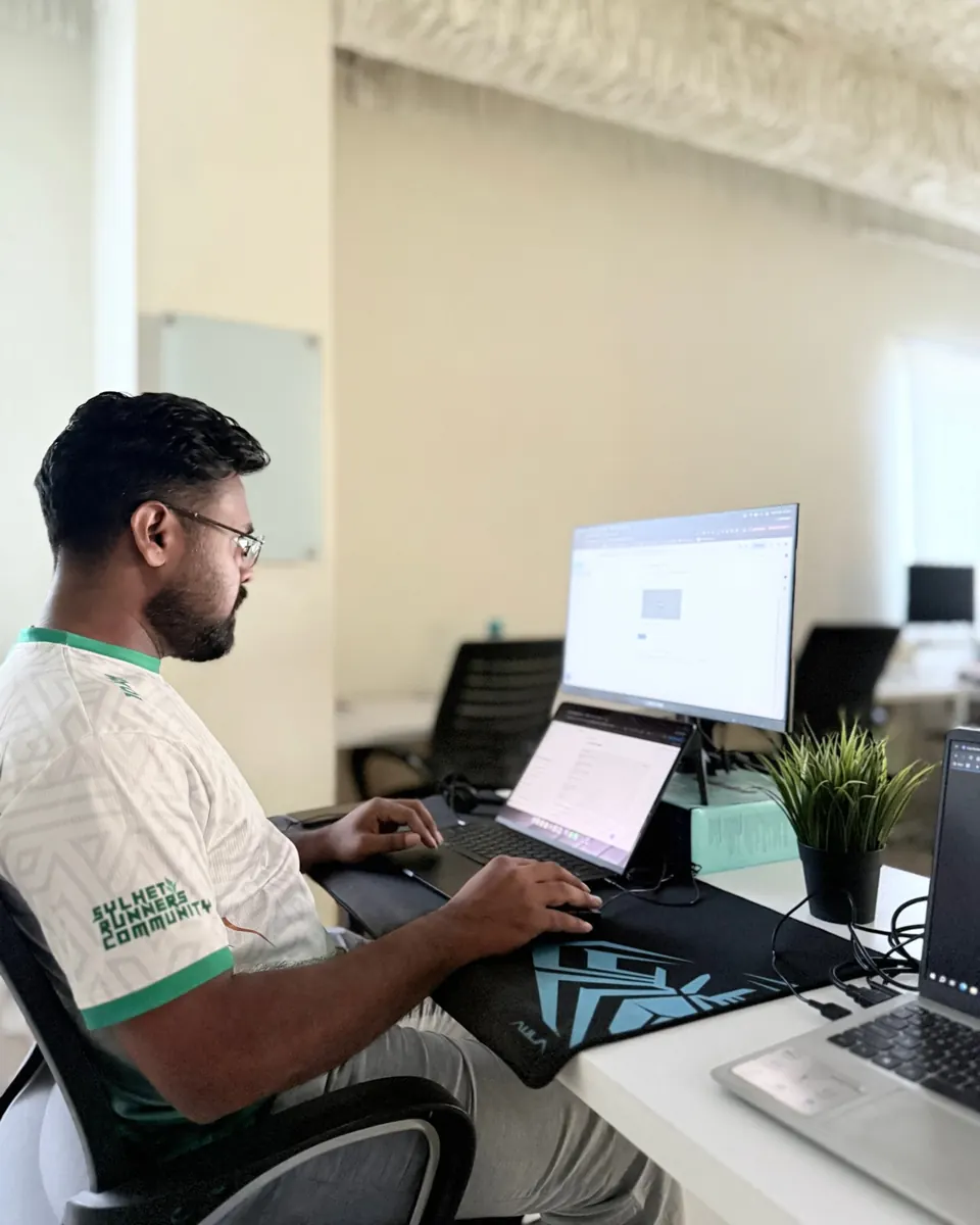 KMR Jibon working across a laptop and monitor, wearing a Sylhet Runners Community shirt
